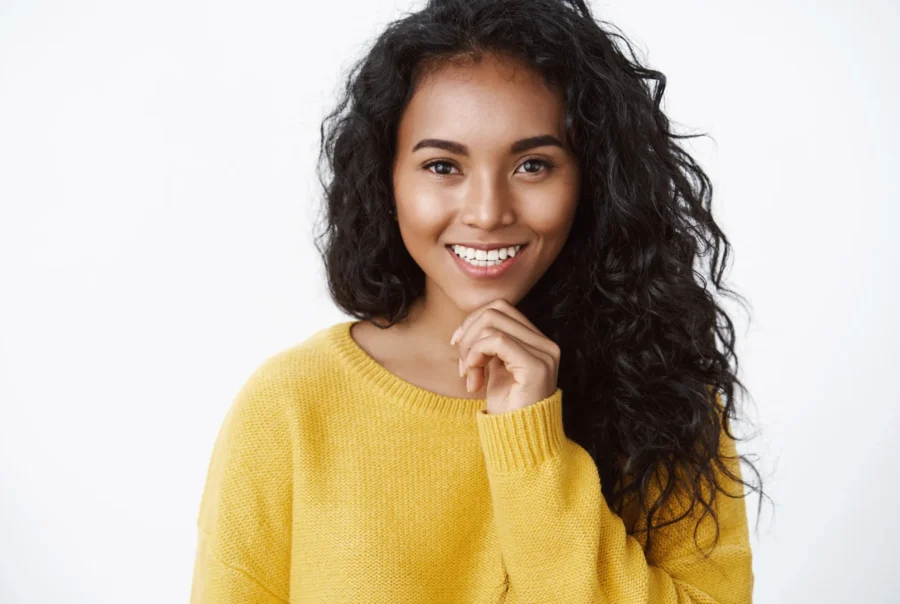 Eyelid Surgery (Blepharoplasty) attractive curly haired woman yellow sweater smiling delighted like what she see as made right choice touch chin thoughtful pondering good idea